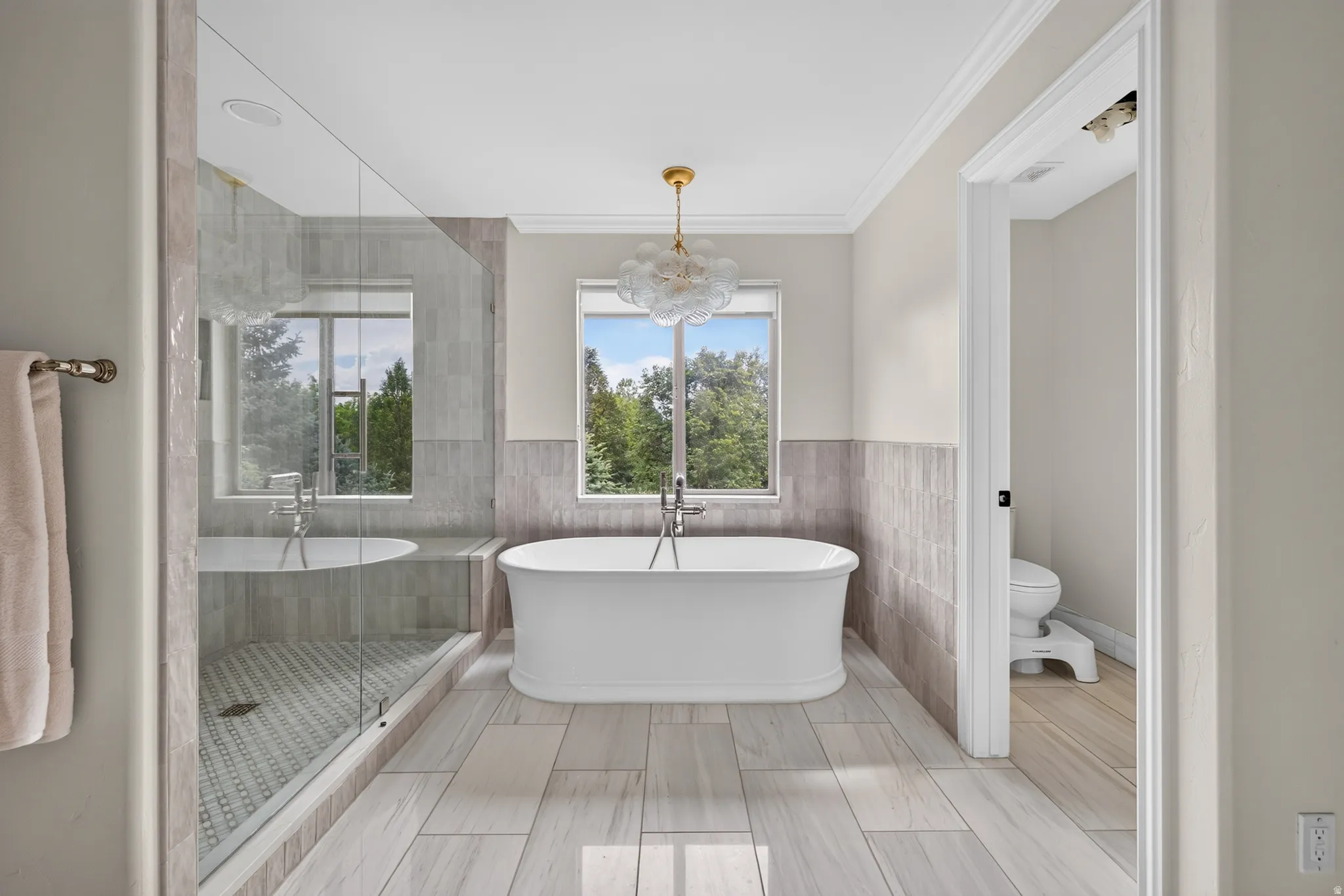 Freestanding soaking tub with a modern chandelier, oversized glass-enclosed shower, wood-finish flooring, and crown molding
