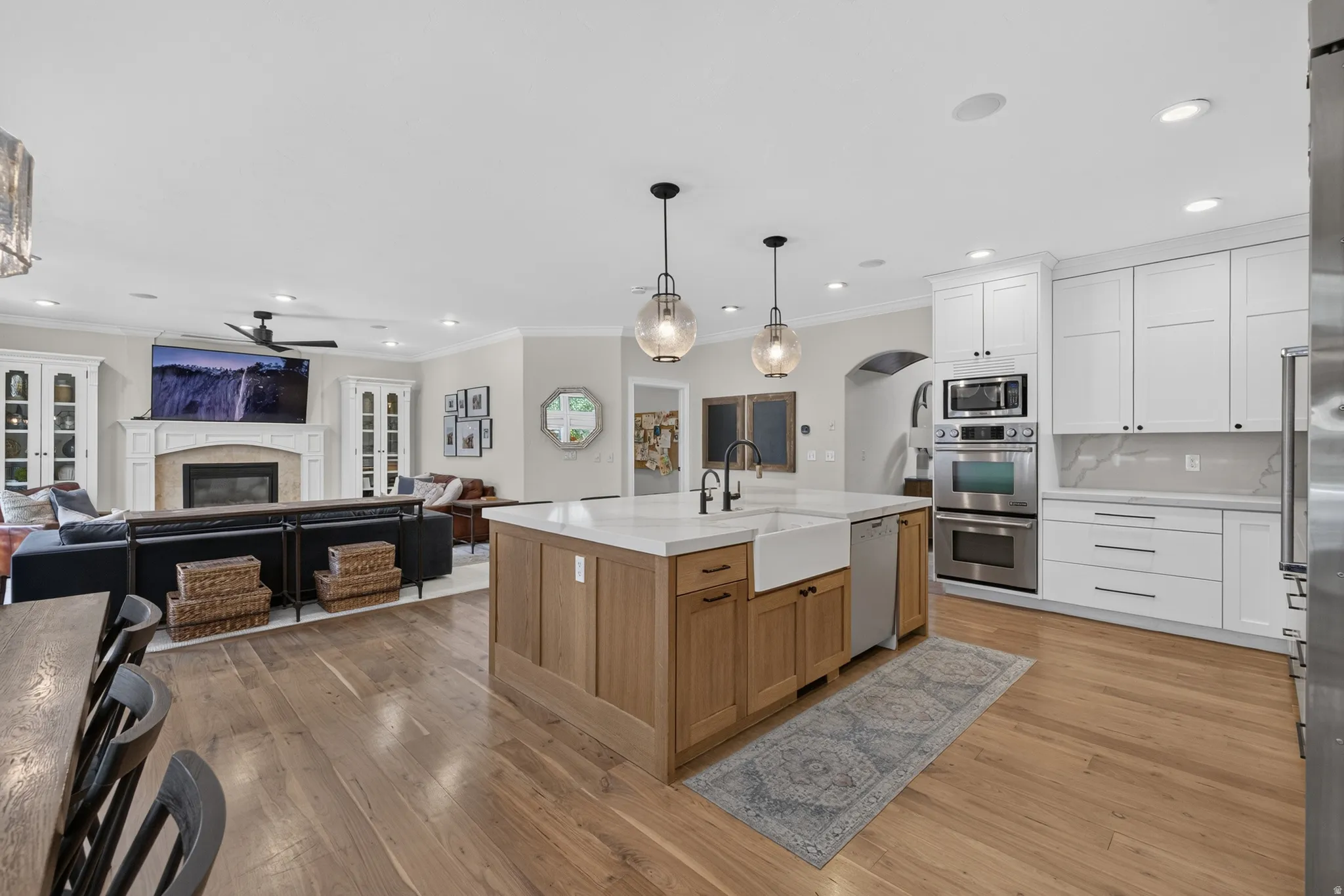 Spacious kitchen featuring a central island with a farmhouse sink, stainless steel appliances, white cabinetry, and wood-finish flooring extending into the living area with a fireplace