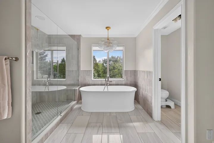 Freestanding soaking tub with a modern chandelier, oversized glass-enclosed shower, wood-finish flooring, and crown molding