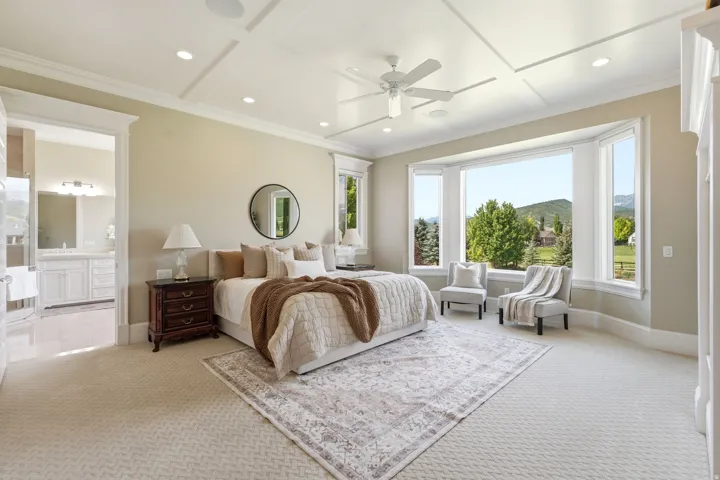 Spacious bedroom featuring a bay window, coffered ceiling with recessed lighting and ceiling fan, neutral carpet flooring, and an open doorway to a bathroom with a double vanity