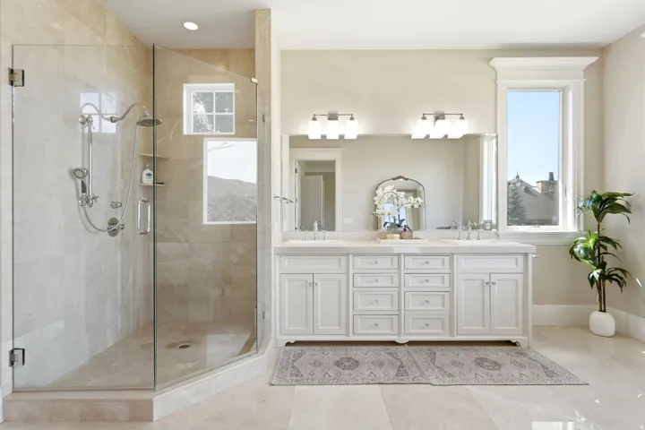 Spacious bathroom featuring a glass-enclosed shower with multiple showerheads, a dual vanity with white cabinetry and undermount sinks, and light-toned tile flooring