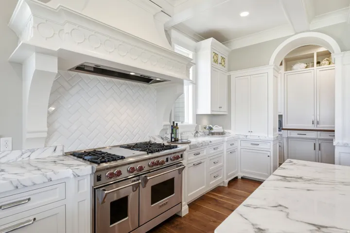 Gourmet kitchen featuring a professional-grade stainless steel range, custom white range hood with decorative molding, white subway tile backsplash in a herringbone pattern, white cabinetry with polished chrome hardware, and marble countertops