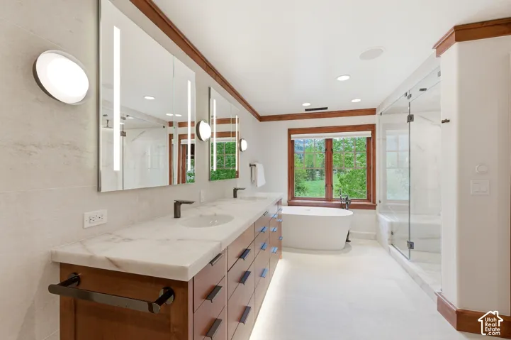 Dual vanity with stone countertops and undermount sinks, contemporary wall-mounted light fixtures, freestanding soaking tub, and a glass-enclosed shower