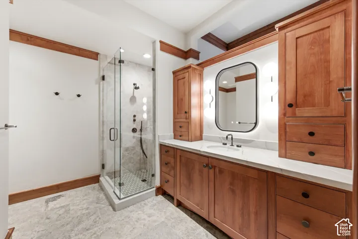 Glass-enclosed shower with multiple spray features, extensive wood cabinetry, vanity with undermount sink, illuminated mirror, and stone flooring