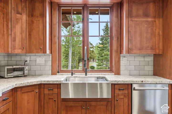 Wood-finish cabinetry with brass hardware, light-toned countertop, subway tile backsplash, stainless steel farmhouse sink, and stainless steel dishwasher