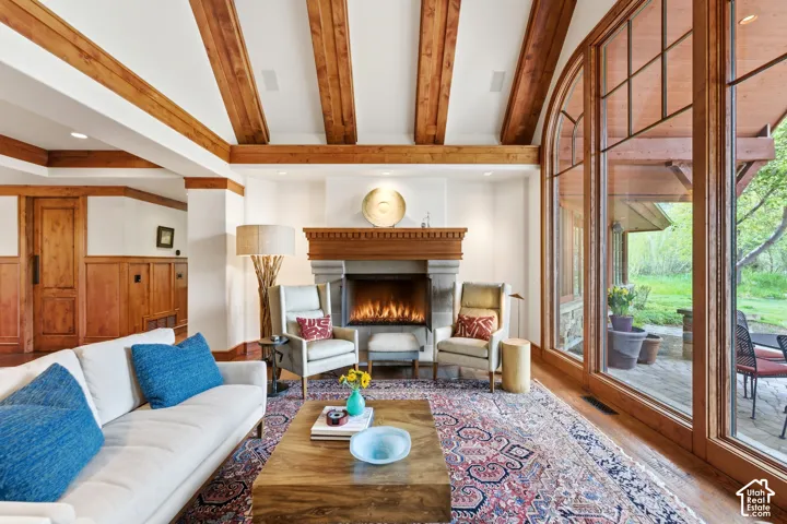 Vaulted ceiling with exposed wood beams, wood-paneled walls, stone fireplace with wood mantel, large arched windows, and a paved outdoor patio