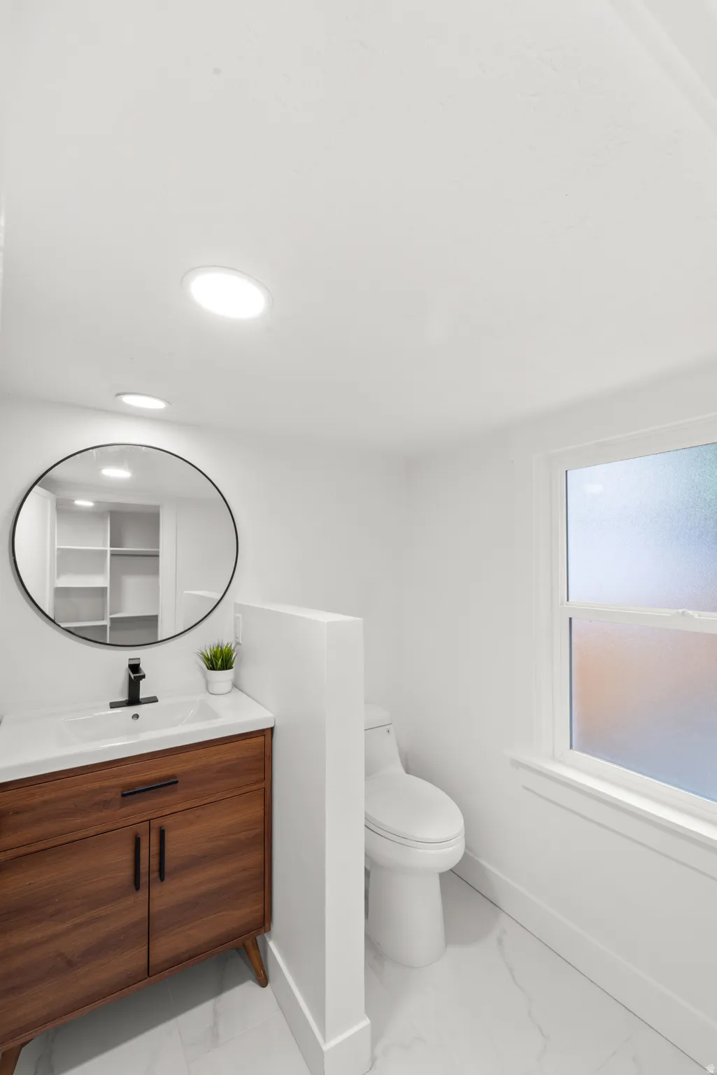 Primary full bathroom featuring a wood-finish vanity with integrated sink, a circular black-framed mirror, recessed lighting, and white marble-look floor tiles