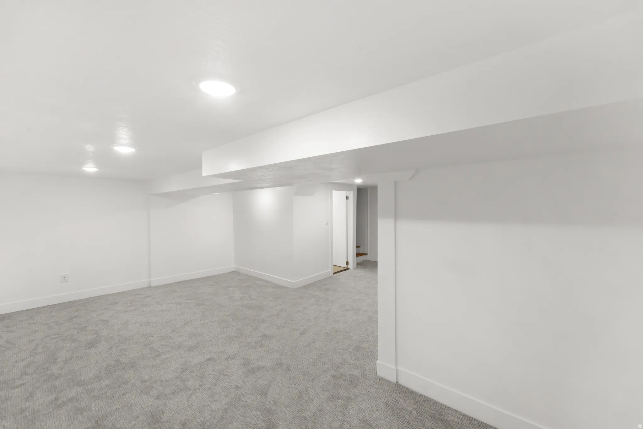 Spacious room featuring light gray carpet, recessed lighting, white walls, and a visible doorway leading to a staircase