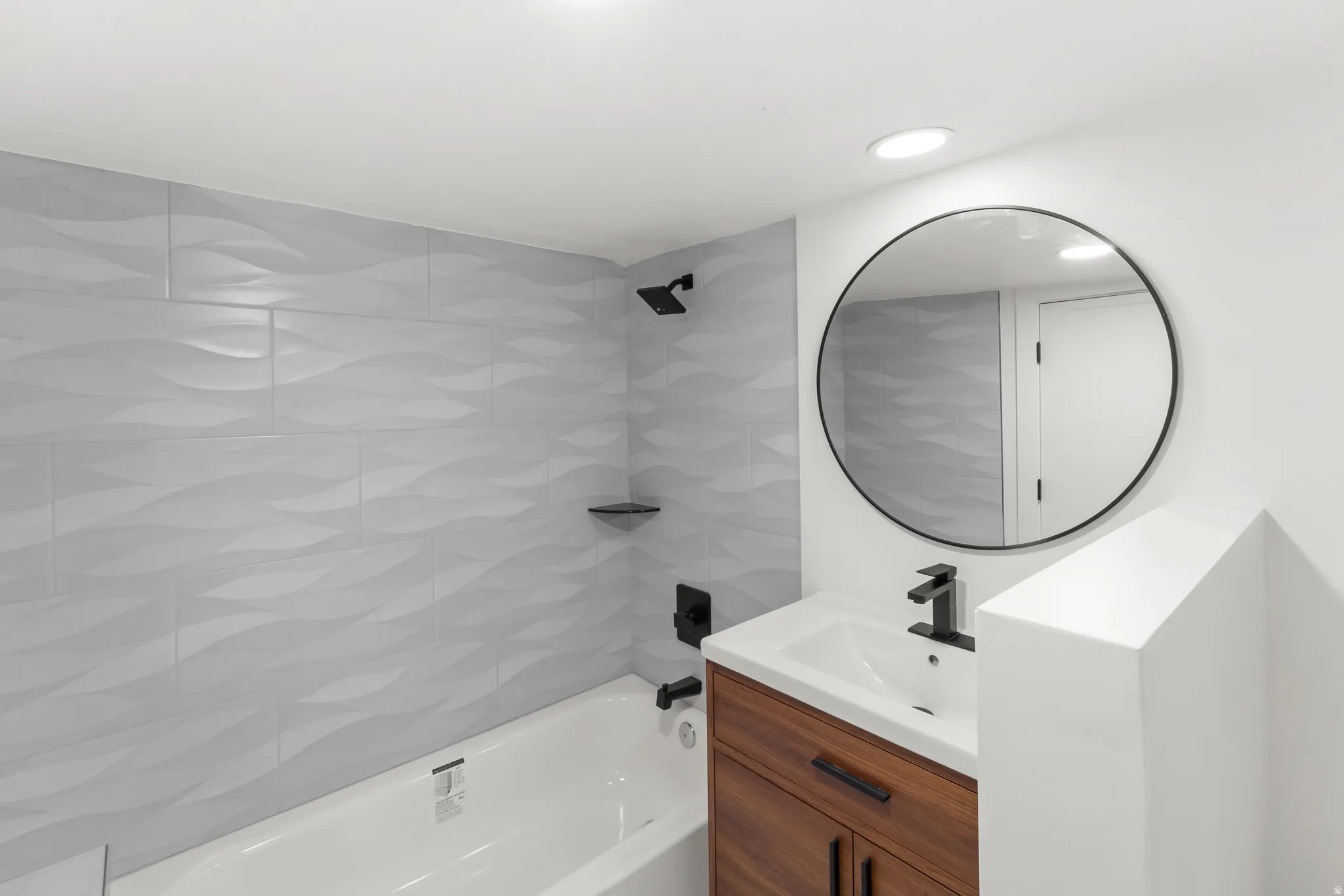 Primary full bathroom featuring a white bathtub with light gray textured tile surround, a dark wood-finish vanity with a white countertop sink, matte black faucet, and a round black-framed mirror