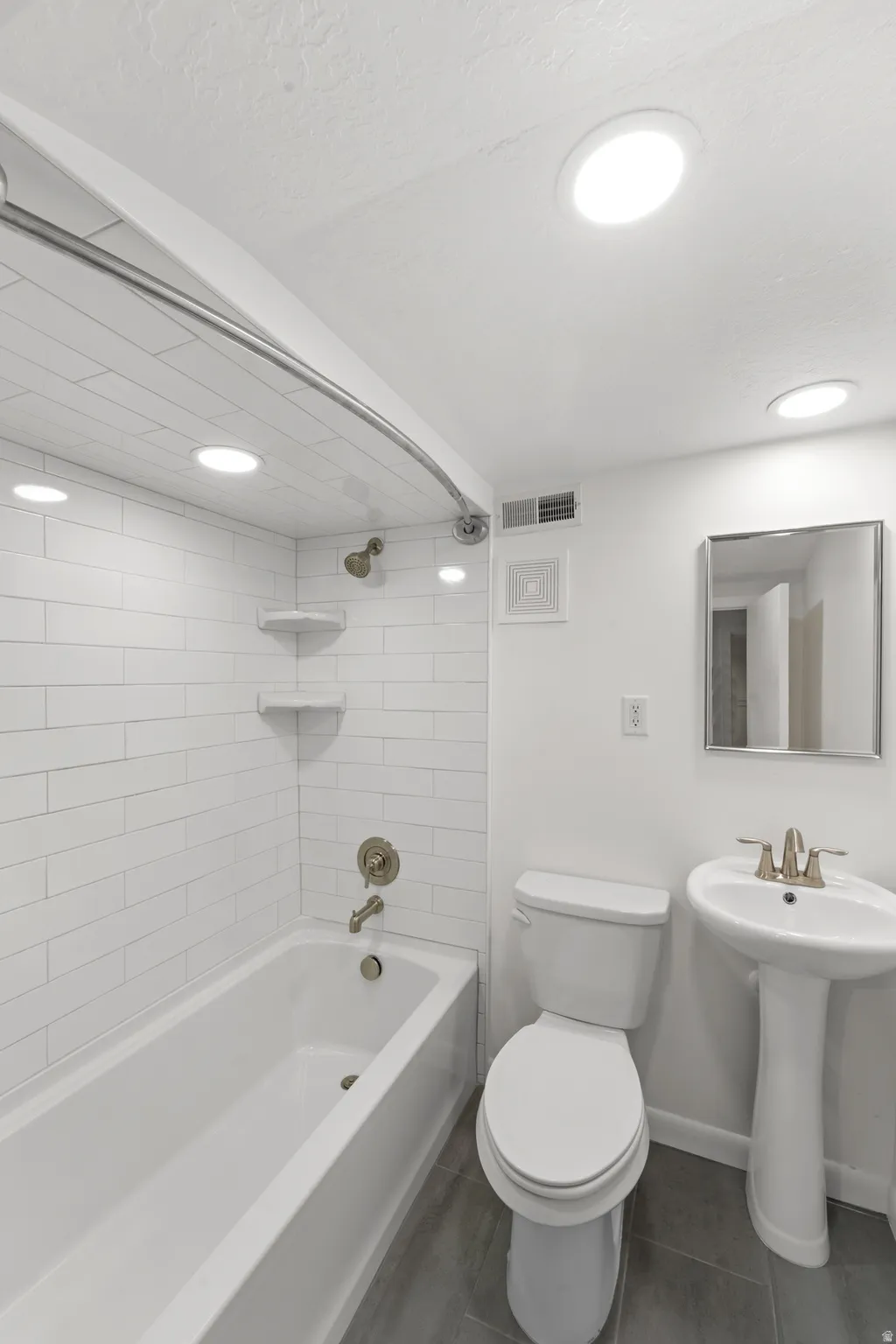 Bathroom featuring a white bathtub with subway tile surround, pedestal sink, toilet, and gray tile flooring