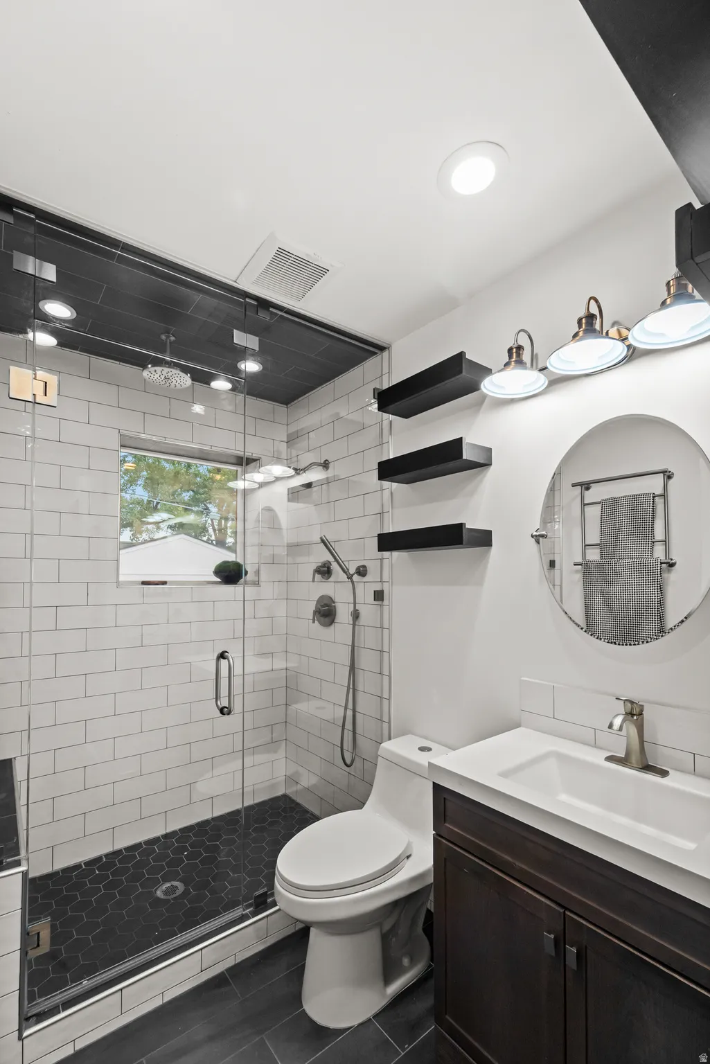 Contemporary bathroom featuring a dark wood-finish vanity with a white integrated sink, a framed glass shower enclosure with white subway tile walls, a dark hexagonal tile shower floor, and dark wood-finish flooring
