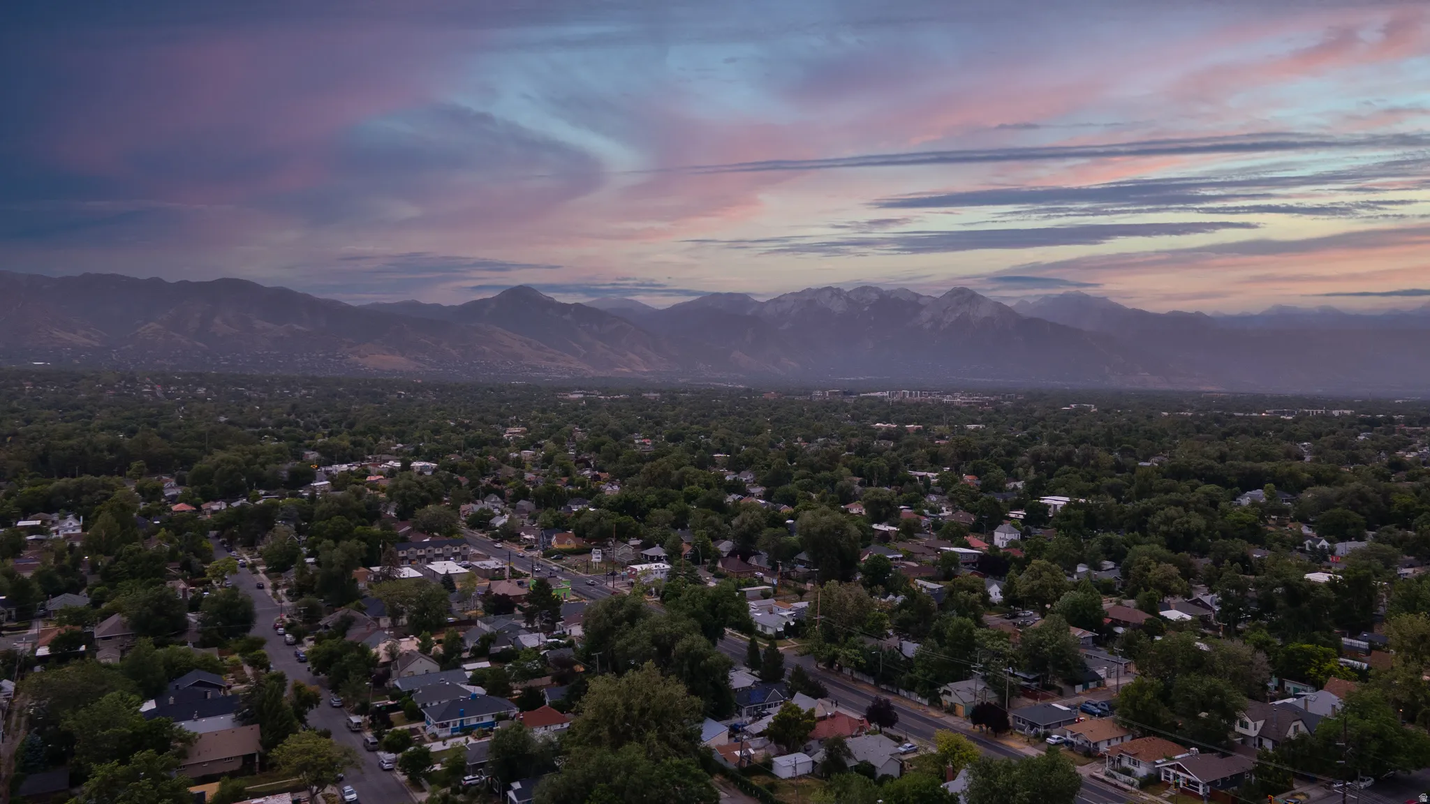 Expansive neighborhood setting featuring a dense canopy of mature trees, residential properties with varied architectural styles, and distant mountains with snow-capped peaks