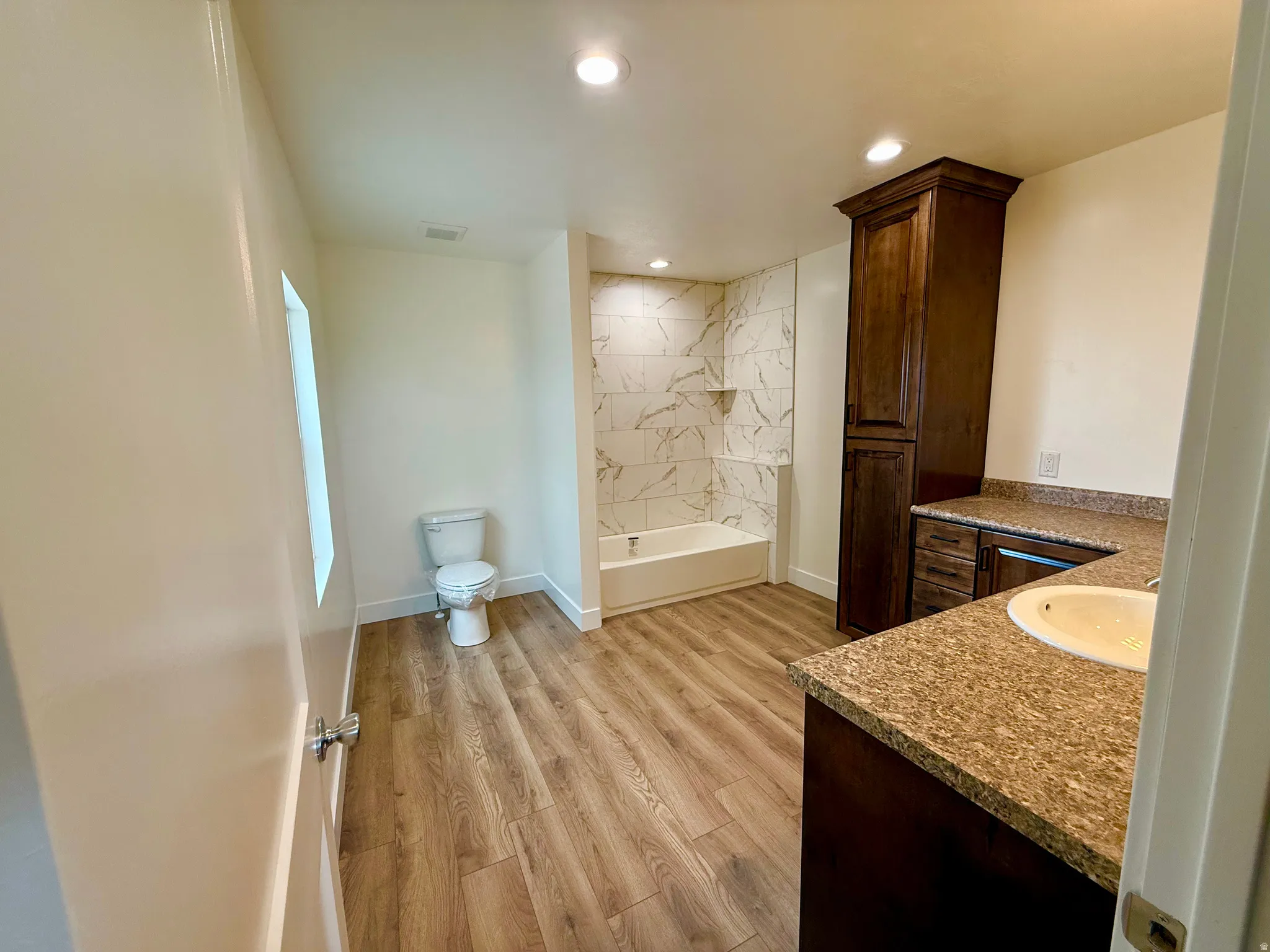 Primary ensuite Bathroom featuring wood-finish flooring, a white tub with white-and-gray patterned tile surround, a dark wood-finish vanity with a light-colored countertop and integrated sink, and recessed lighting