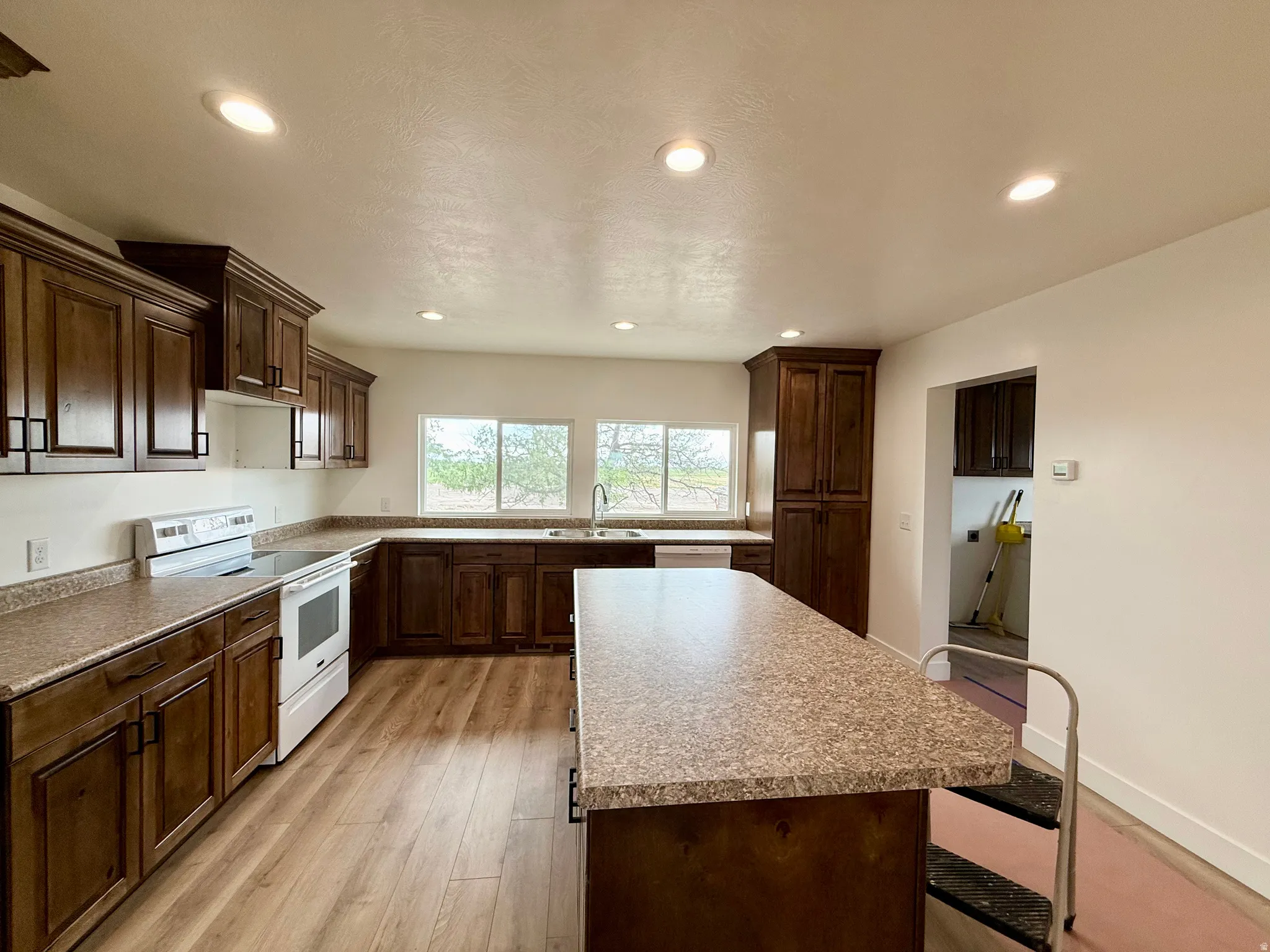 Spacious kitchen featuring wood-finish flooring, recessed lighting, ample dark wood cabinetry with matte black hardware, and an expansive island with a speckled countertop