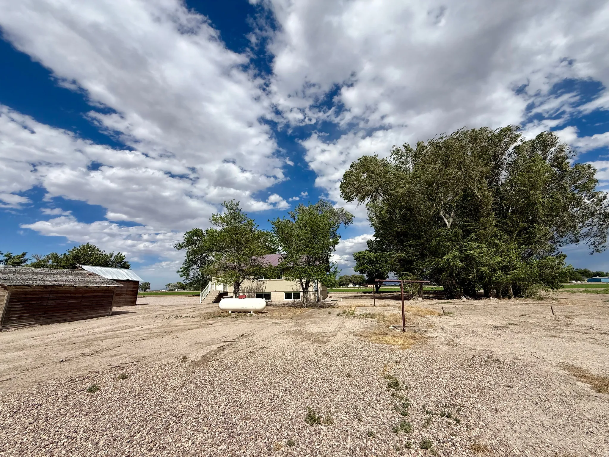 Expansive gravel lot featuring a large outbuilding with a metal roof, a white residential structure with exterior stairs, and multiple mature trees
