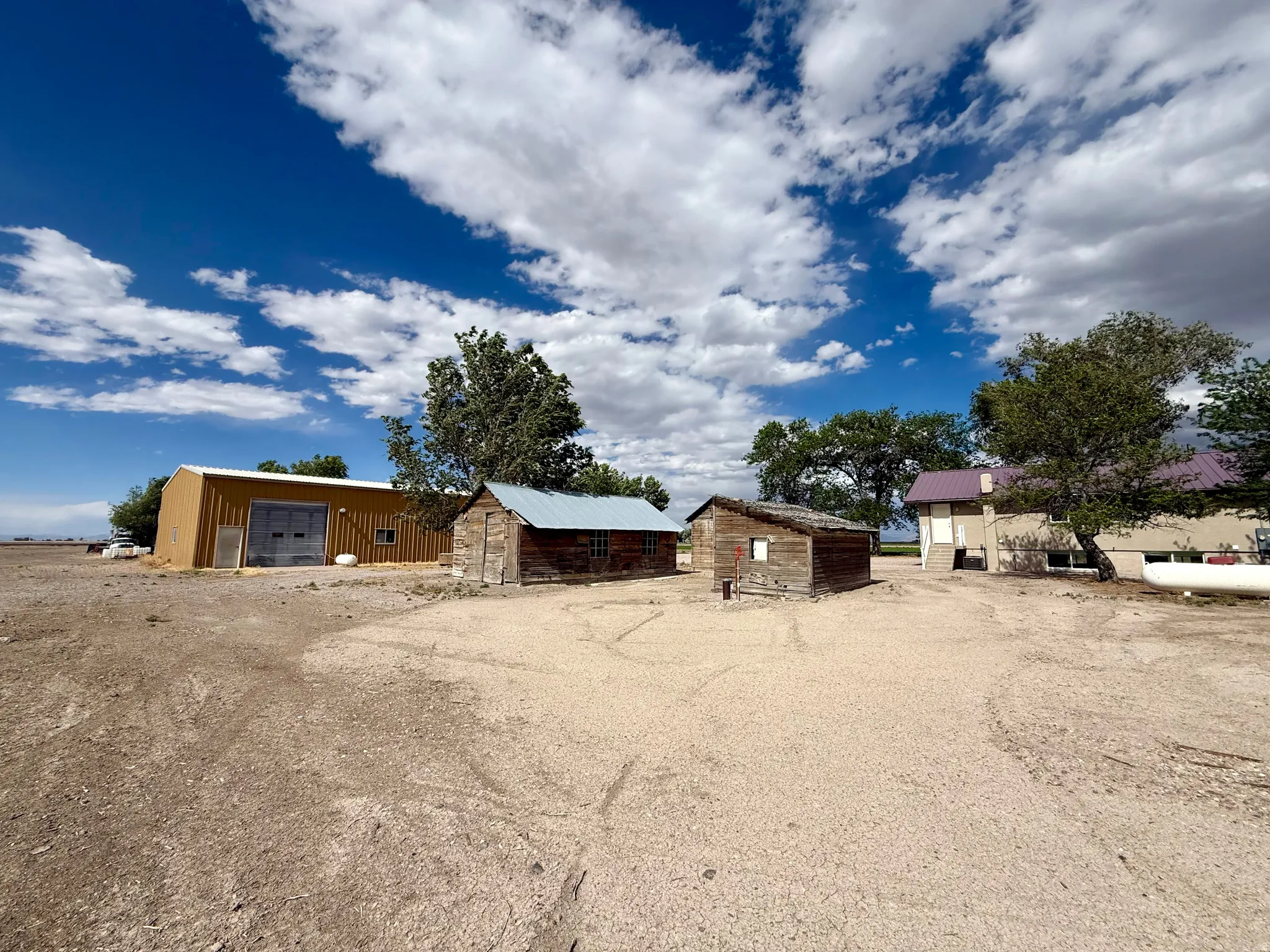 Expansive property featuring a large tan metal building with dual bay doors, two rustic wooden outbuildings with metal roofs, and a multi-level structure with a light-colored exterio