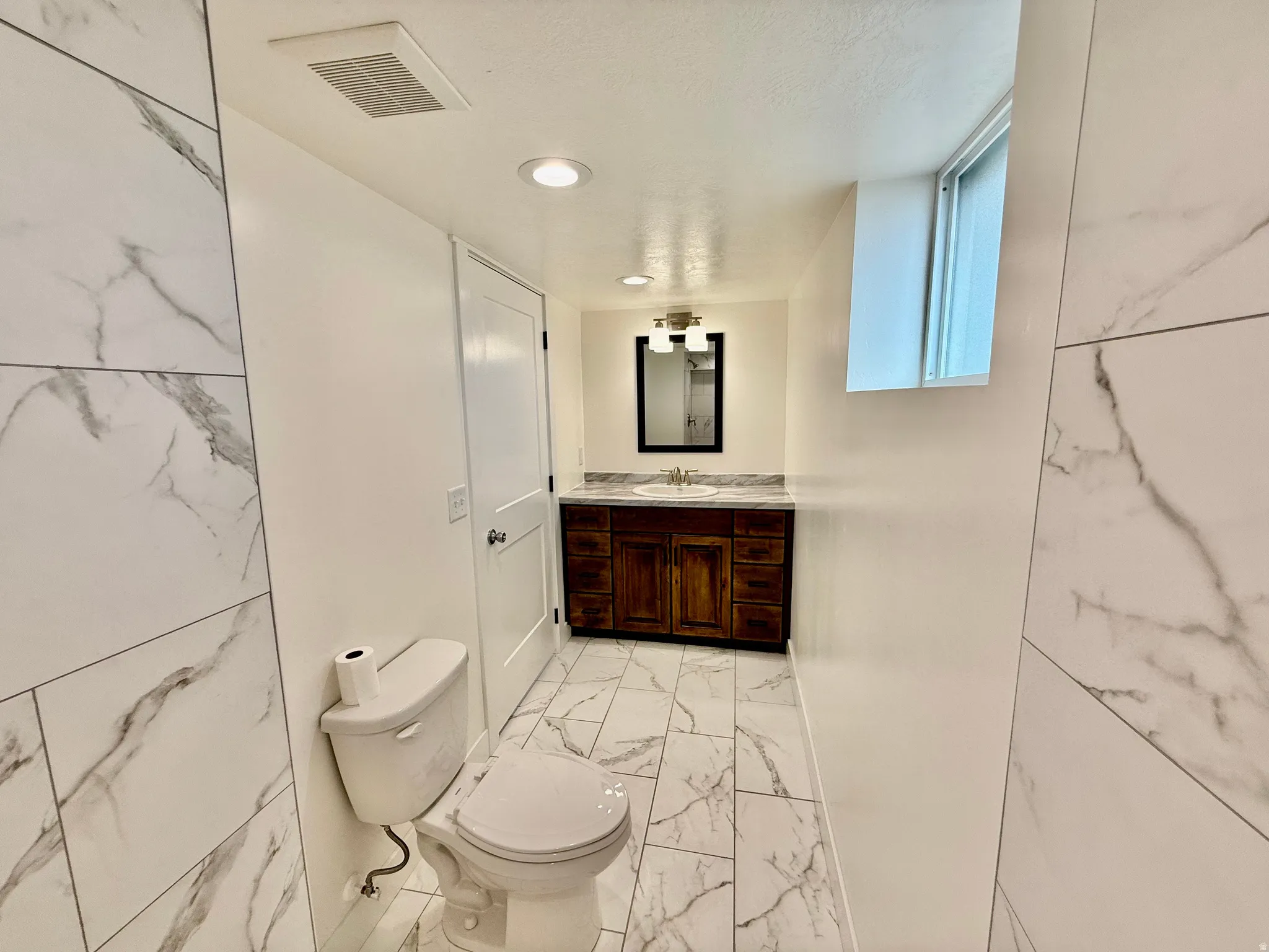 Bathroom featuring large-format tile flooring and shower surround with marble-look veining, a dark wood-finish vanity with a light-colored countertop, and a contemporary light fixture