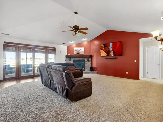 Spacious living area featuring a vaulted ceiling with a ceiling fan, a fireplace with a stone surround and wood mantle, and wood-finish sliding glass doors