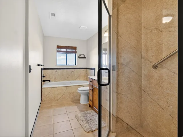 Spacious bathroom featuring a glass-enclosed shower with stone-look wall panels, a built-in tub with stone-look surround and black fixtures, a wood-finish vanity with a countertop and undermount sink, and tiled flooring