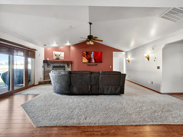 Spacious living area featuring a vaulted ceiling with a ceiling fan, a fireplace with a stone surround and wood mantel, large glass sliding doors, wood-finish flooring, and wall sconces