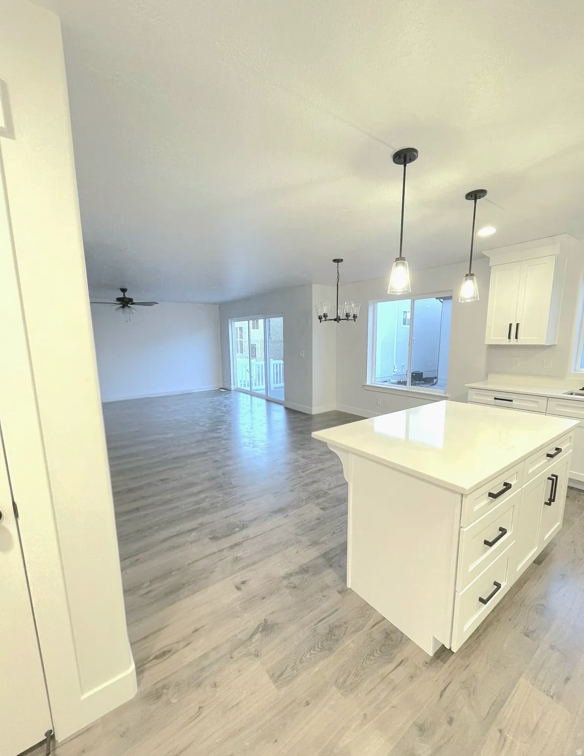 Open-concept living area featuring light wood-finish flooring, a kitchen island with a light-toned countertop, white cabinetry with dark hardware, and pendant lighting