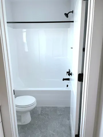 Bathroom featuring a white tub/shower combination with matte black fixtures, white toilet, and gray stone-finish flooring