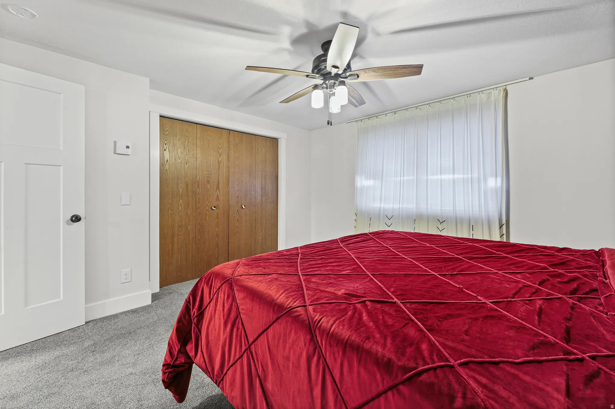 Bedroom featuring soft gray carpeting, a multi-paneled white door with a black knob, a ceiling fan with integrated lighting, and a window with sheer white coverings