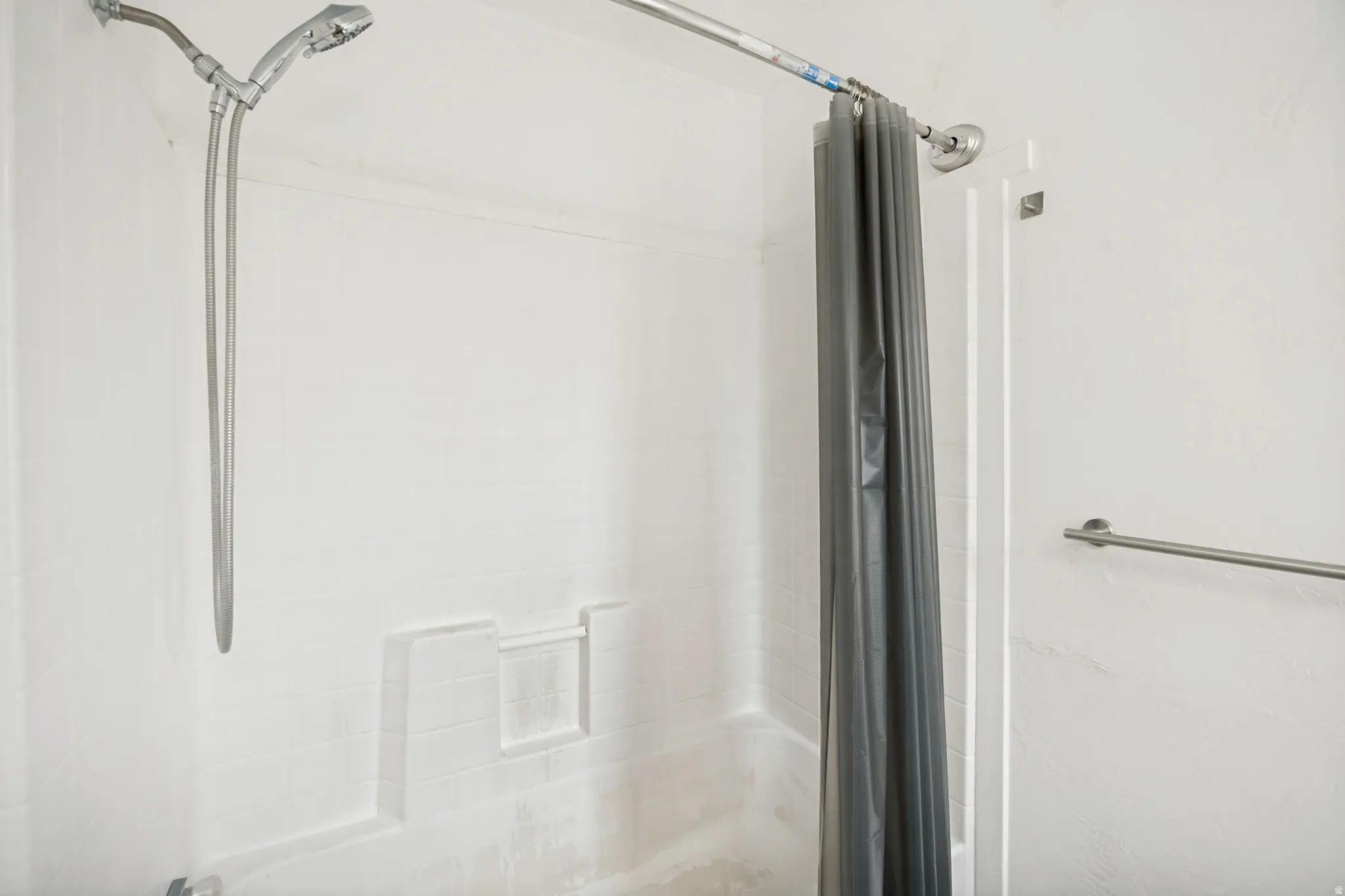 Bathtub and shower combination featuring a white tiled surround, chrome shower head, chrome handheld sprayer, built-in shelving, and a chrome towel bar