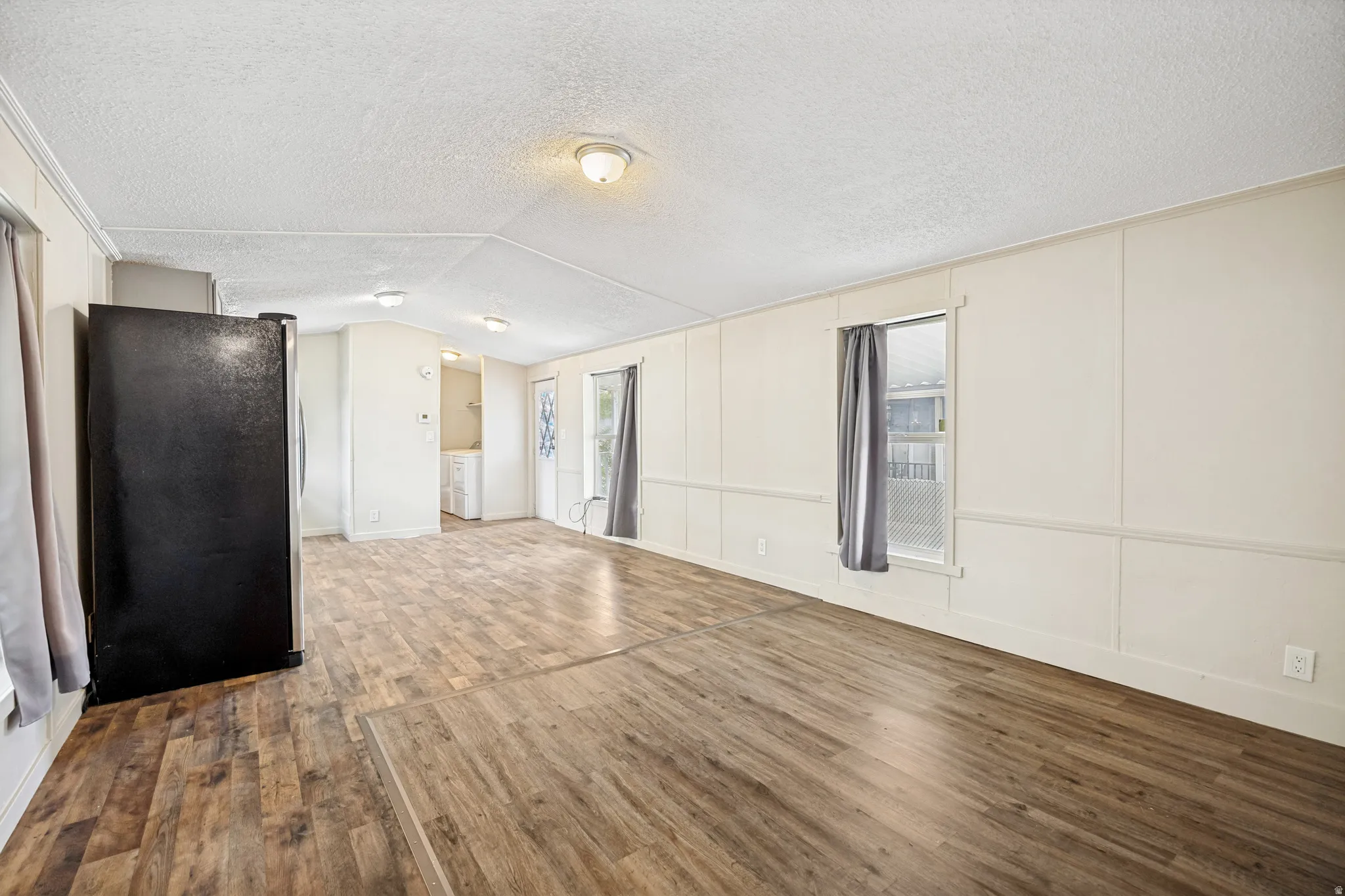 Expansive living area featuring wood-finish flooring, light-toned paneled walls, multiple windows with drapes, and flush-mount ceiling lighting