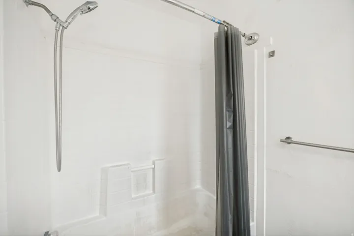 Bathtub and shower combination featuring a white tiled surround, chrome shower head, chrome handheld sprayer, built-in shelving, and a chrome towel bar