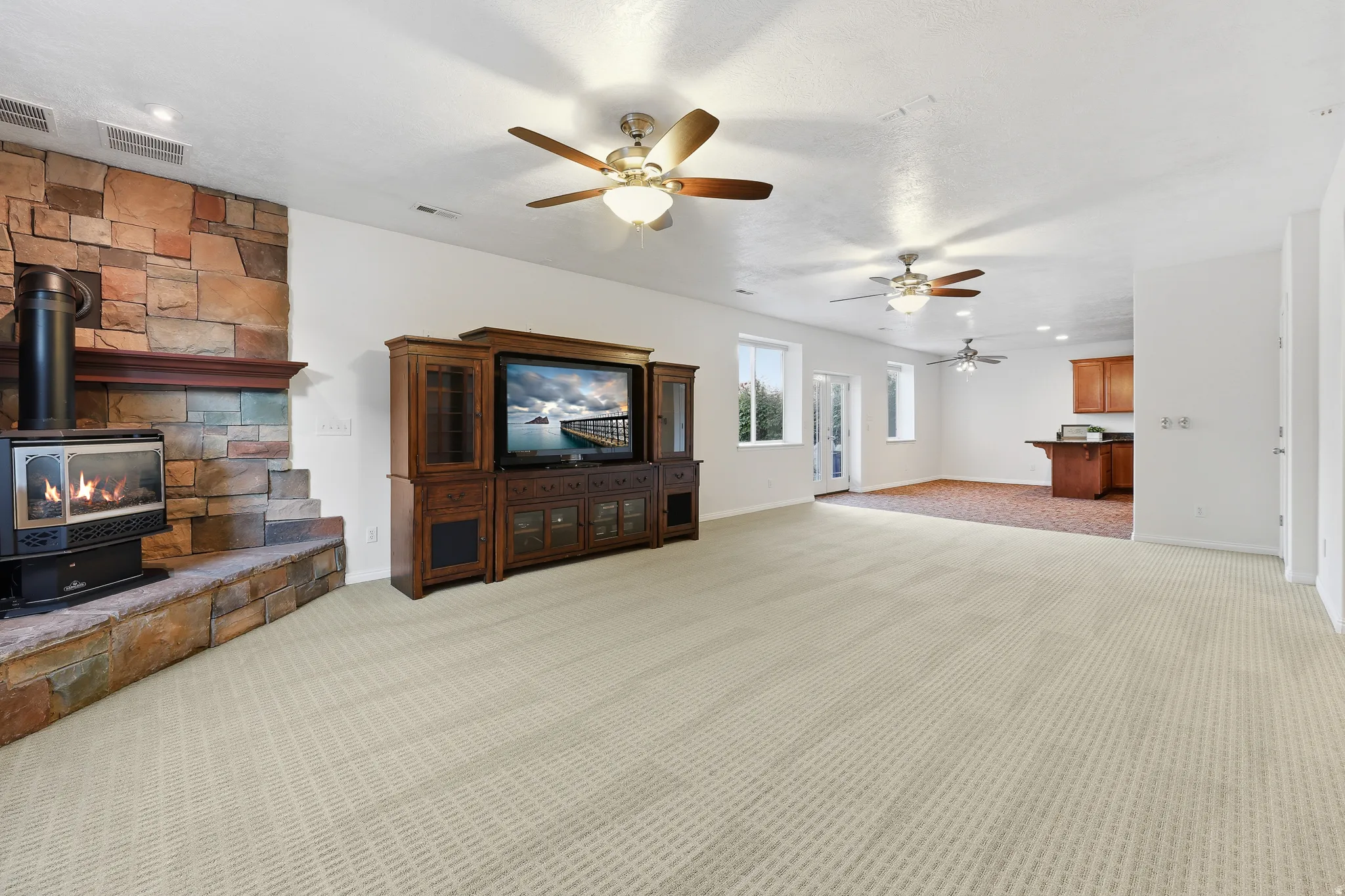 Spacious room featuring a stone-clad fireplace with hearth, multiple ceiling fans, light-toned patterned carpeting, and recessed lighting