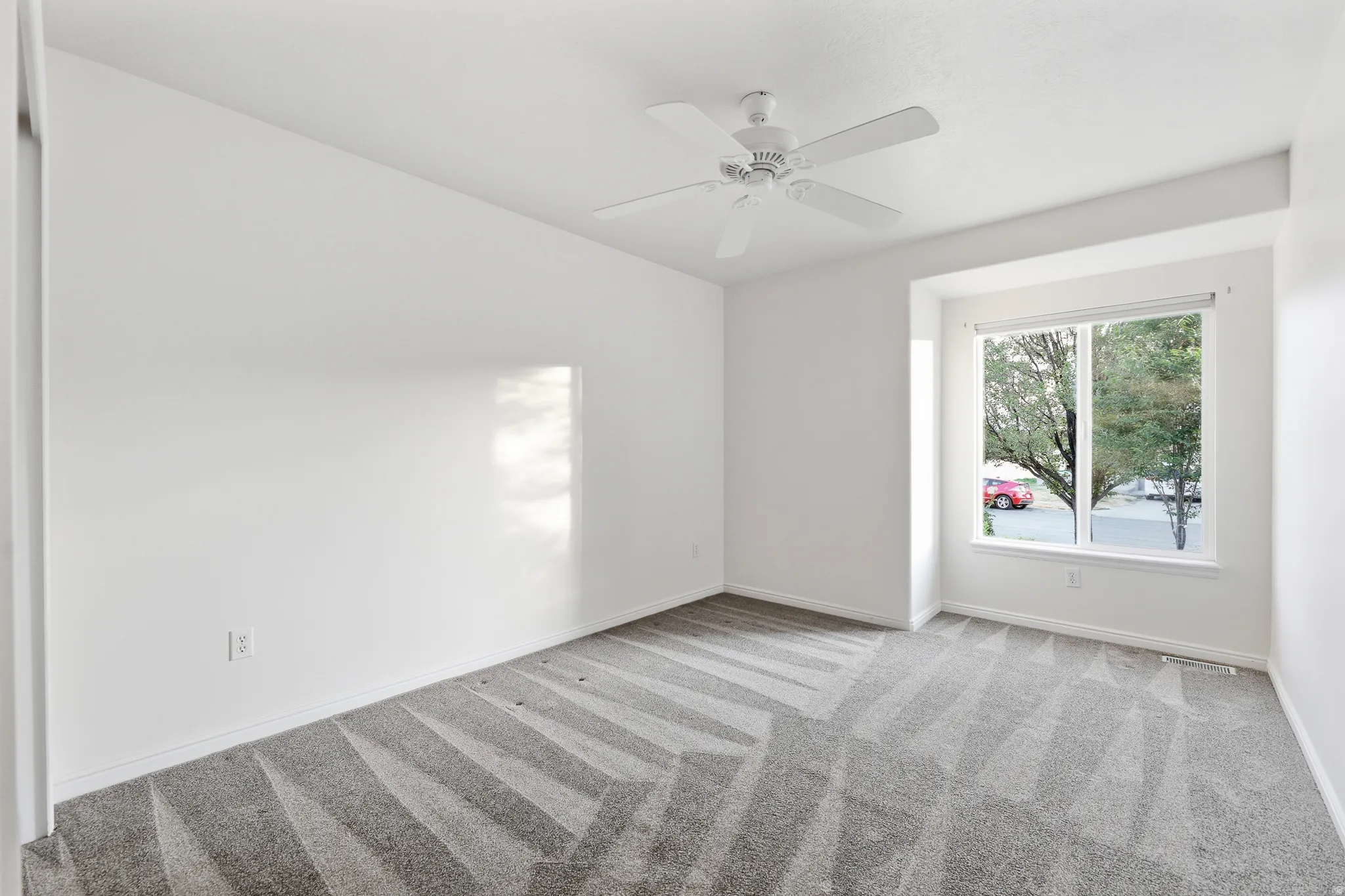 Bright bedroom featuring a large window, neutral carpeting with geometric patterning, and a ceiling fan