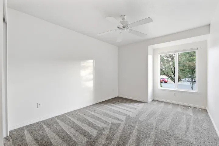 Bright bedroom featuring a large window, neutral carpeting with geometric patterning, and a ceiling fan