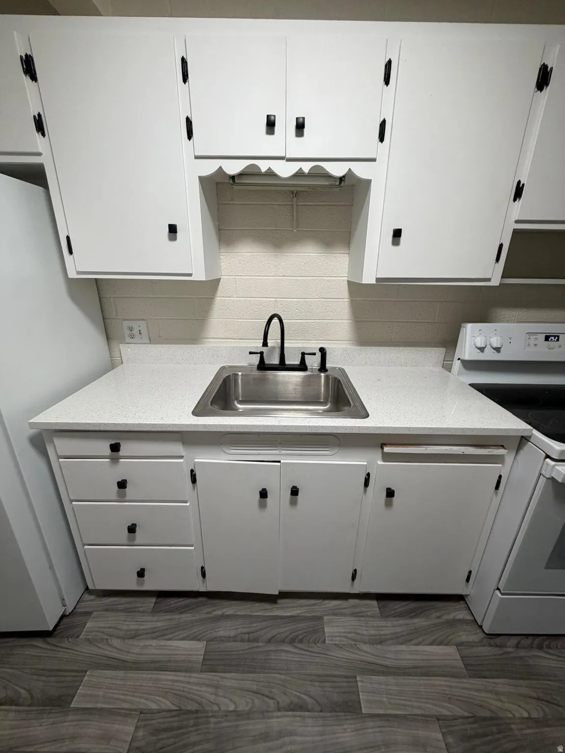 Kitchen featuring white cabinetry with black hardware, a stainless steel sink with a matte black faucet, light-toned countertops, and wood-finish flooring