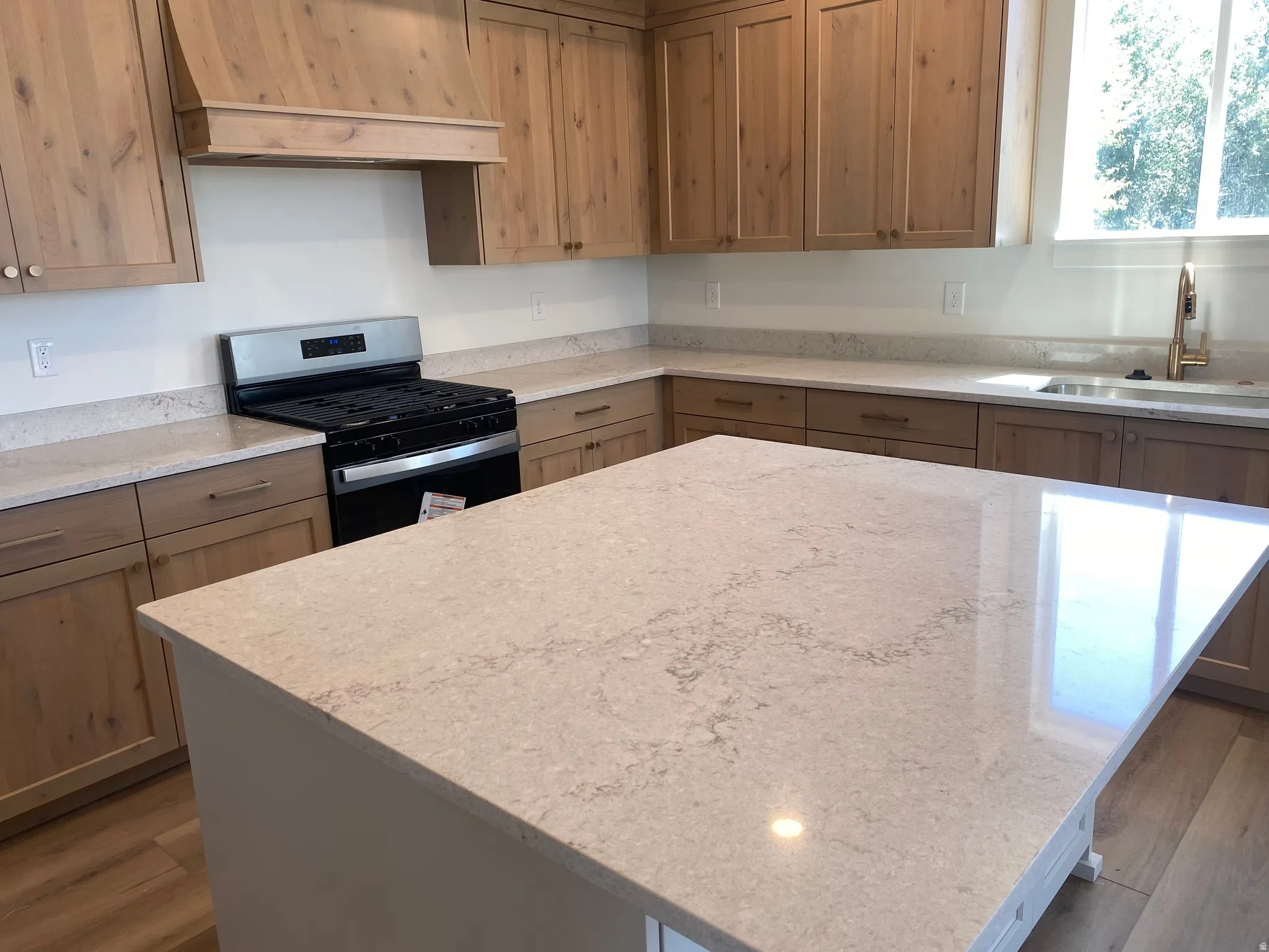 Kitchen featuring a large central island with a light-toned countertop, wood-finish flooring, natural wood cabinetry, a stainless steel range, and a window above the sink