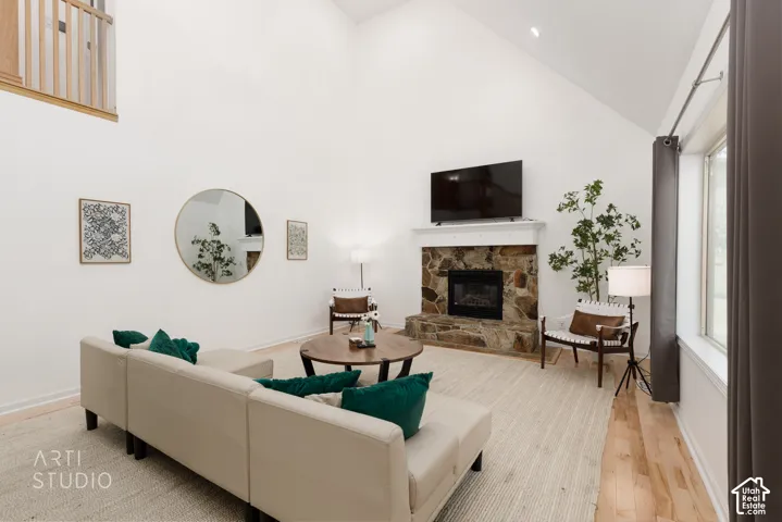Vaulted ceiling living area featuring a stone-clad fireplace with a white mantel, wood-finish flooring, and a large window with dark drapes