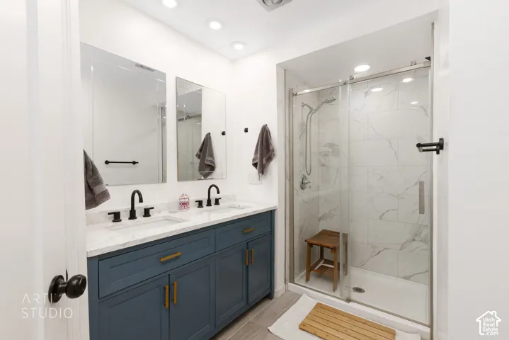 Bathroom featuring a dual vanity with a blue cabinetry and light-toned countertop, two framed mirrors, recessed lighting, and a glass-enclosed shower with tiled walls