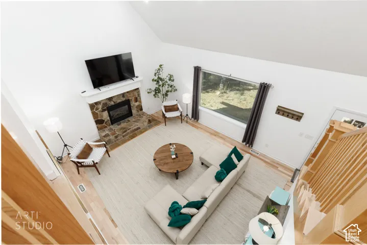 Expansive living area featuring a vaulted ceiling, stone fireplace with a white mantel, wide window, light wood-finish flooring, and a natural wood staircase