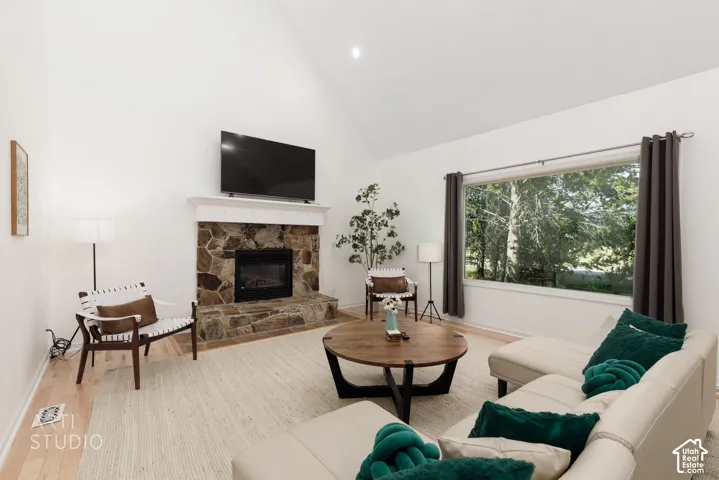 Vaulted ceiling living area featuring a stone-surround fireplace with hearth, a wood-finish floor, and a large picture window with outdoor scenery