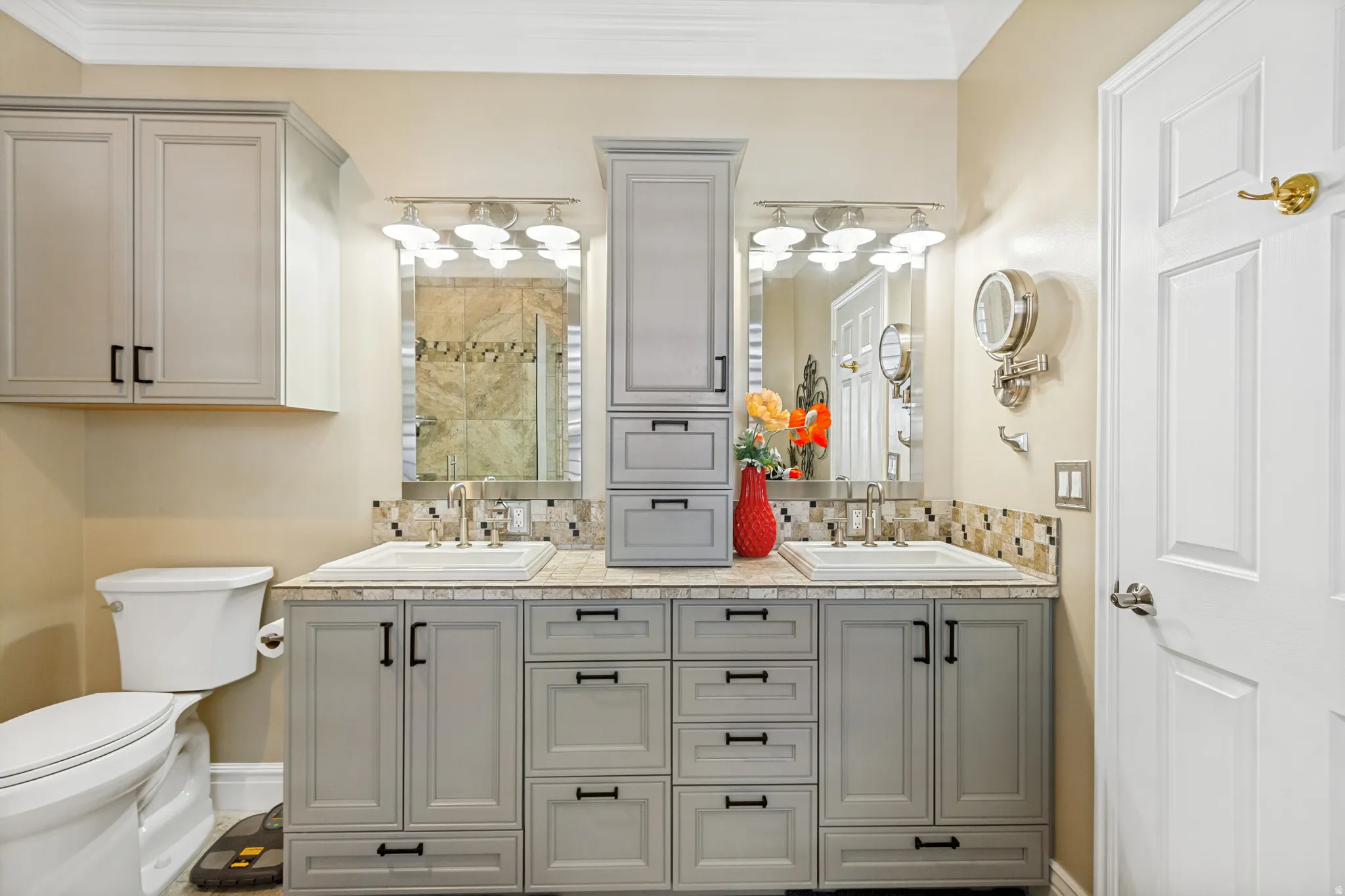 Dual vanity with light gray shaker cabinetry, integrated sinks, a mosaic tile backsplash, and coordinating wall-mounted storage