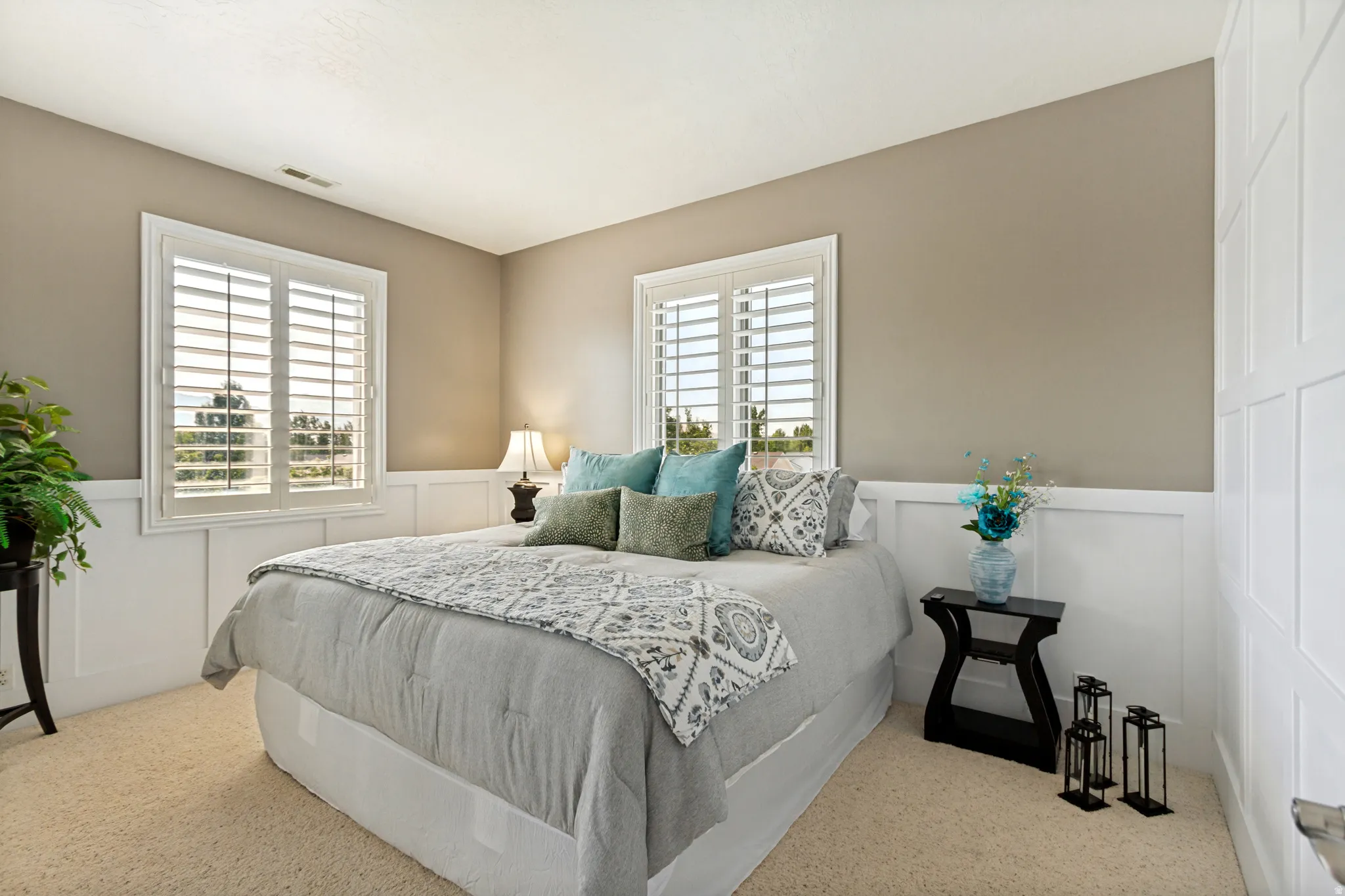 Neutral-toned bedroom featuring white paneling, two windows with plantation shutters, and light-colored carpet flooring