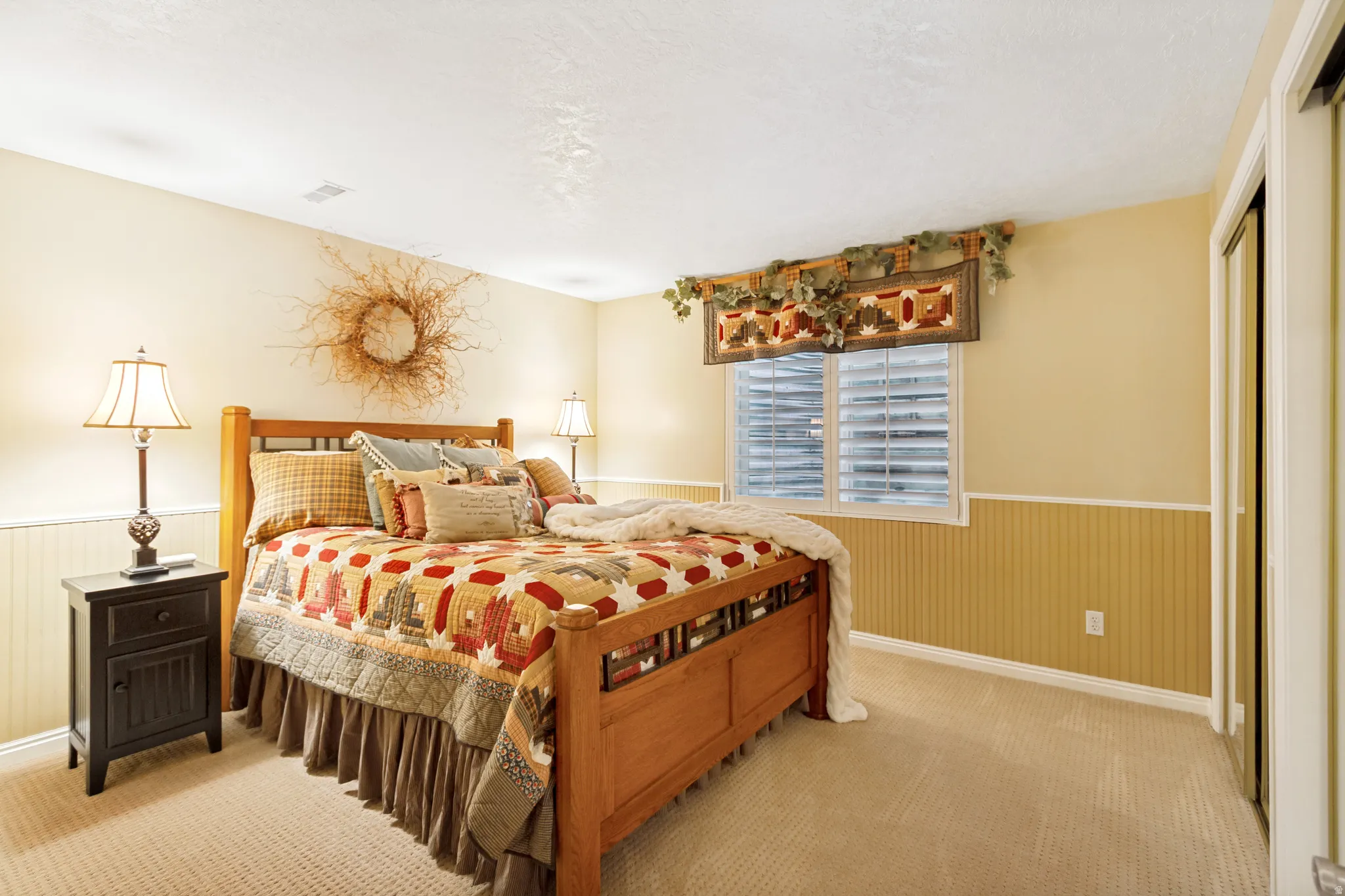 Bedroom featuring wainscoting, neutral carpet flooring, wood-framed window with plantation shutters, and a mirrored closet door