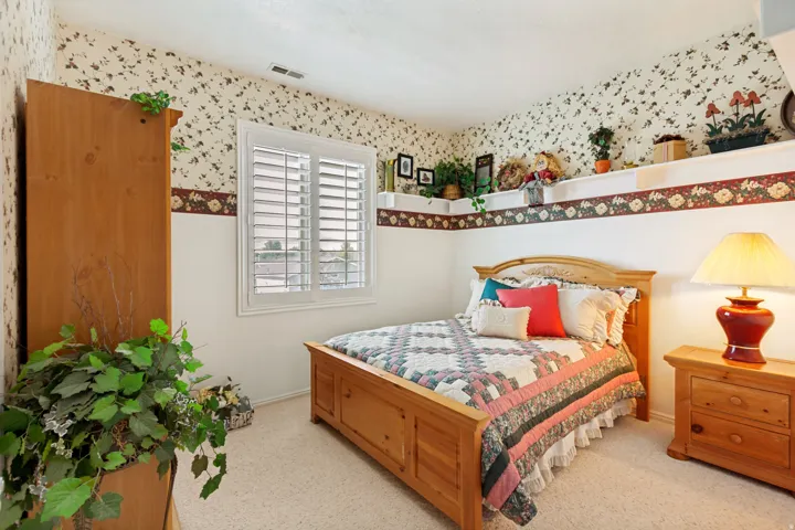 Bedroom featuring a window with white louvered shutters, wall shelving, and patterned wallpaper