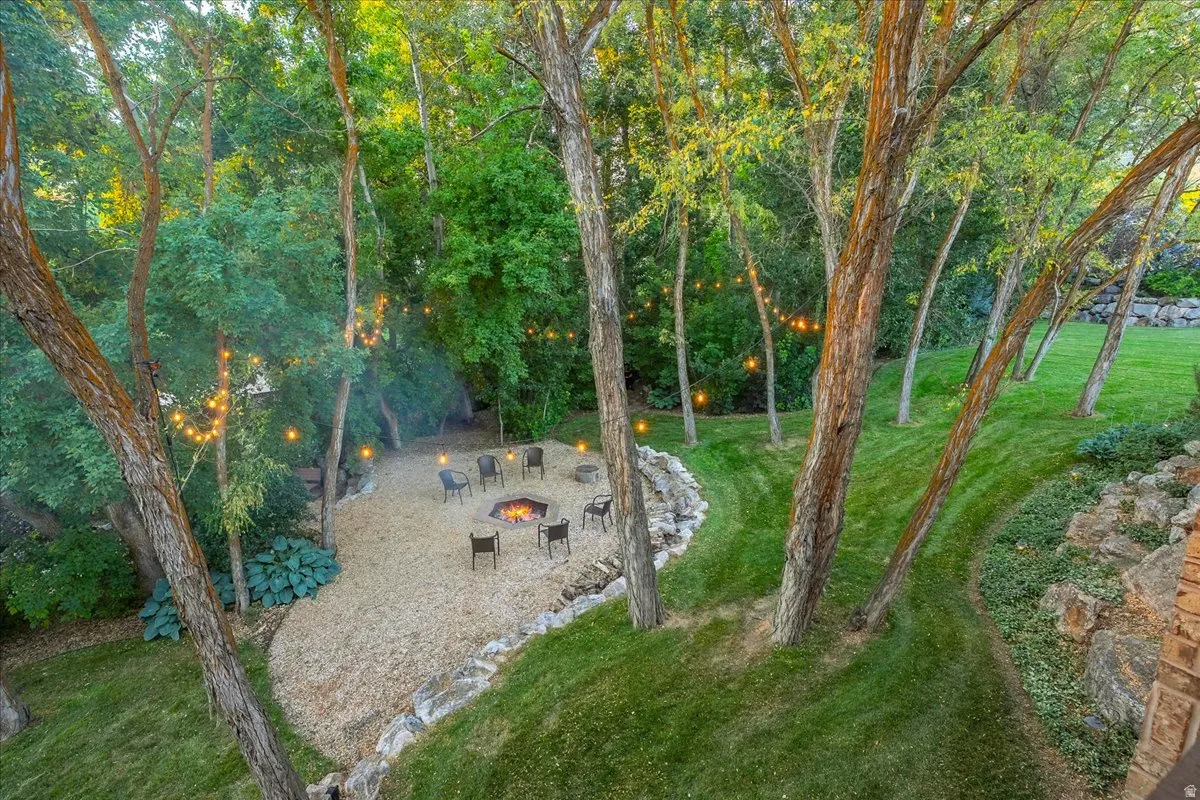 Expansive outdoor entertaining area featuring a gravel fire pit with built-in seating, surrounded by mature trees, string lighting, and manicured lawns