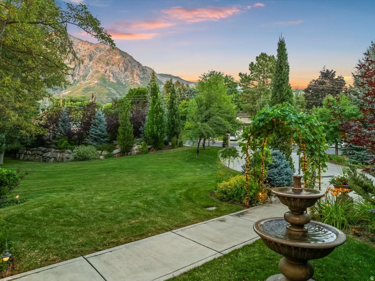 Estate front yard featuring a tiered water fountain, mature landscaping, concrete pathway, decorative archway, and mountain backdrop
