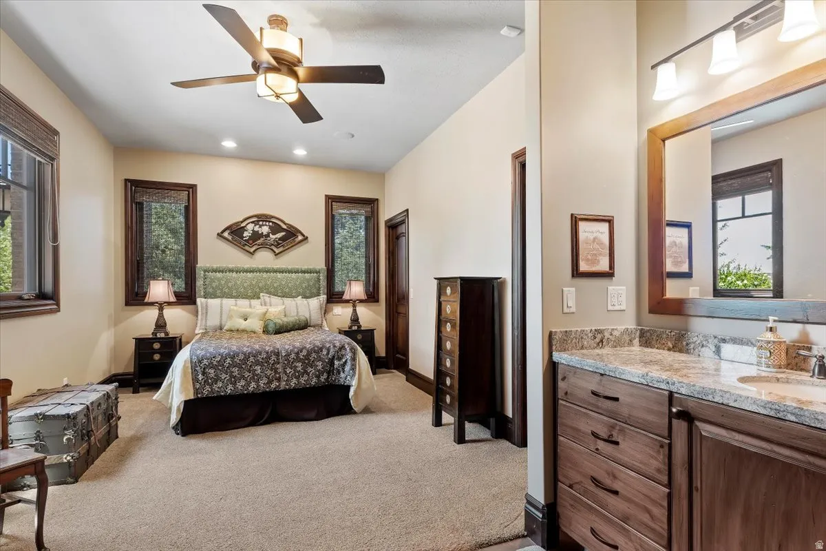 Carpeted bedroom with multiple dark-framed windows, a ceiling fan, recessed lighting, and an attached vanity featuring a granite countertop, undermount sink, and wood-finish cabinetry