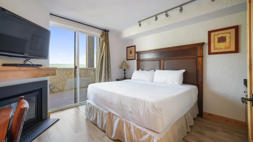 Bedroom featuring a spacious bed with a dark wood headboard, wood-finish flooring, a built-in fireplace with a natural wood mantel, track lighting, and direct access to a private stone-accented balcony through sliding glass doors