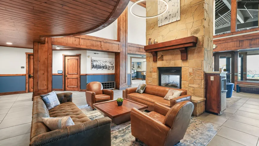 Spacious lounge featuring a stone fireplace with a wood mantle, exposed wood beams, wood-paneled ceilings, and light tile flooring