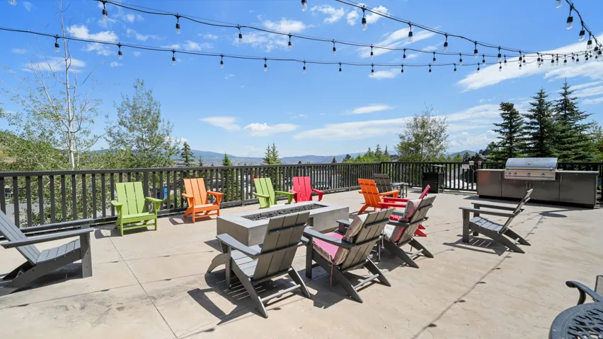 Spacious outdoor patio with string lighting, a gas fire pit, and an outdoor kitchen featuring a built-in grill, all surrounded by a dark wood railing