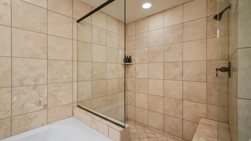 Tiled shower with a clear glass enclosure, integrated bench seating, built-in shelving, recessed lighting, and a rainfall shower head with bronze fixtures
